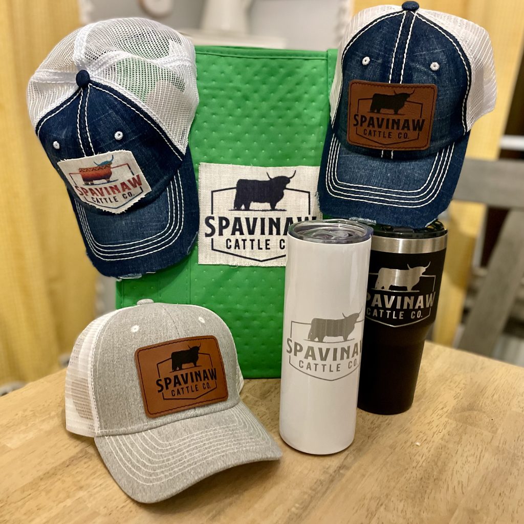 Branded Spavinaw Cattle Co trucker hats, insulated tumblers, and a green bag on display.