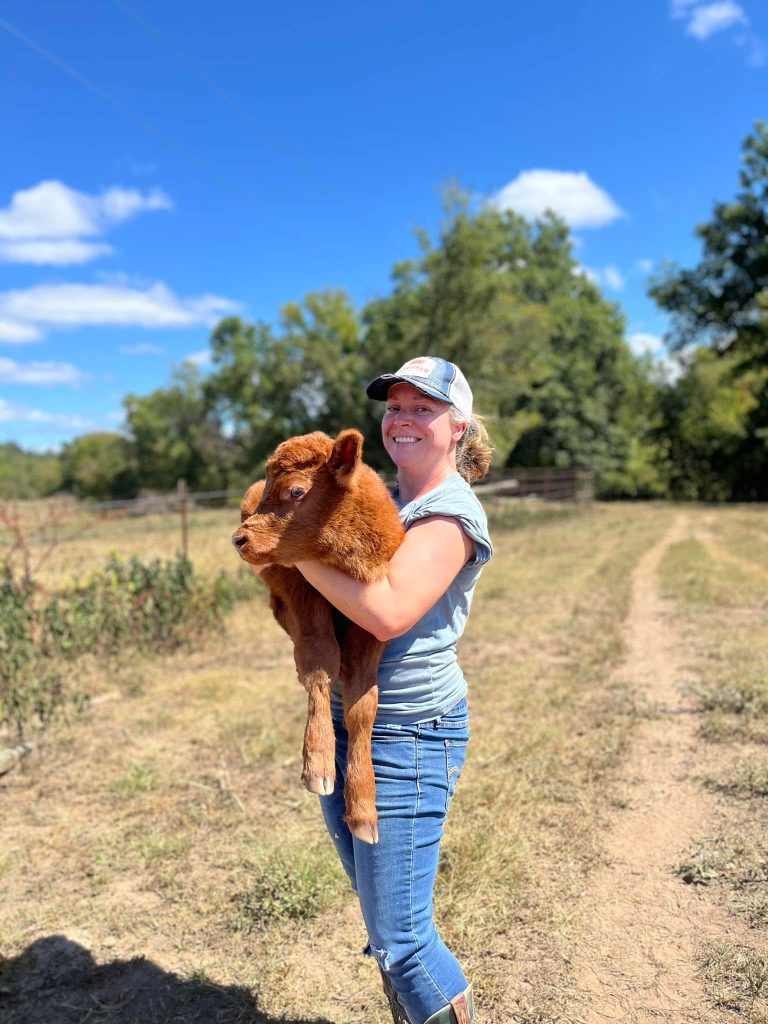 A smiling woman in a farm cap holding a reddish-brown calf in the pasture.