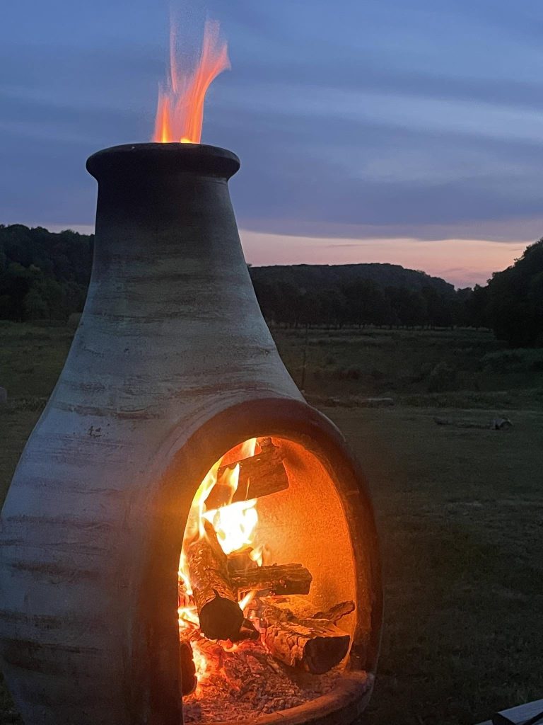 A glowing chiminea with pasture and a pink-and-blue evening sky behind it.