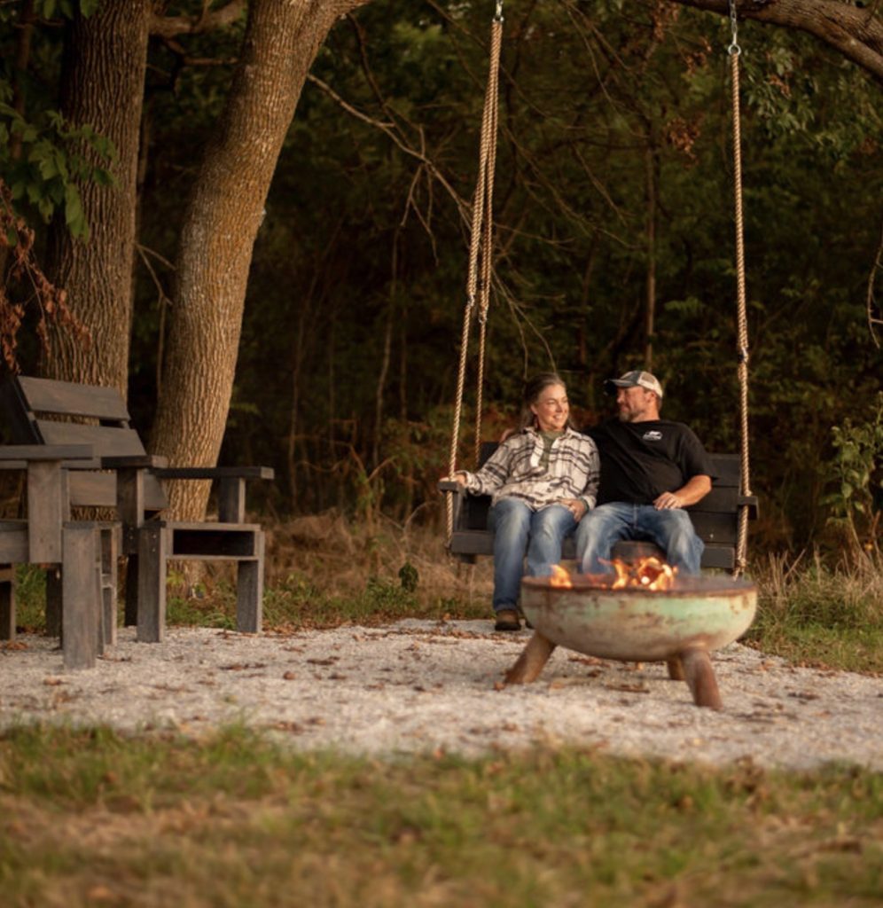 Two adults sitting together on a wooden swing behind an outdoor firepit.