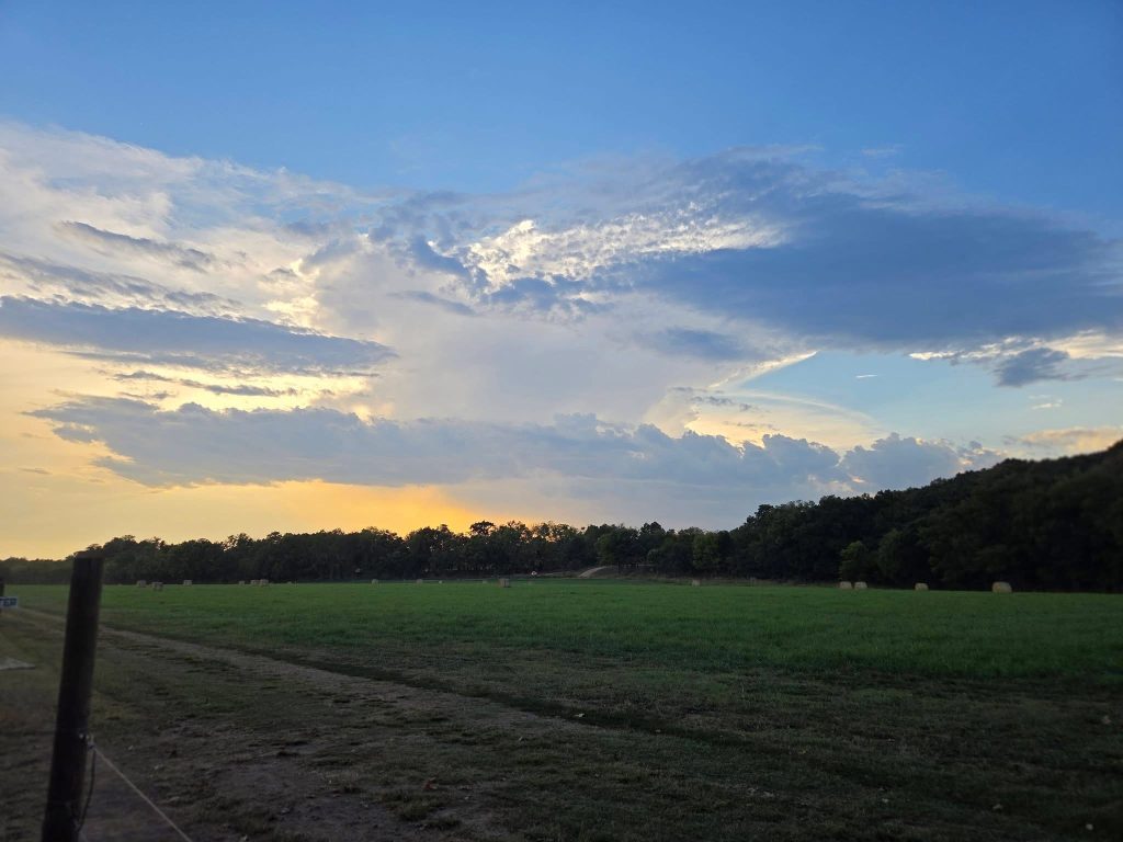 Clouds glowing in the evening light above a broad green field dotted with hay bales.