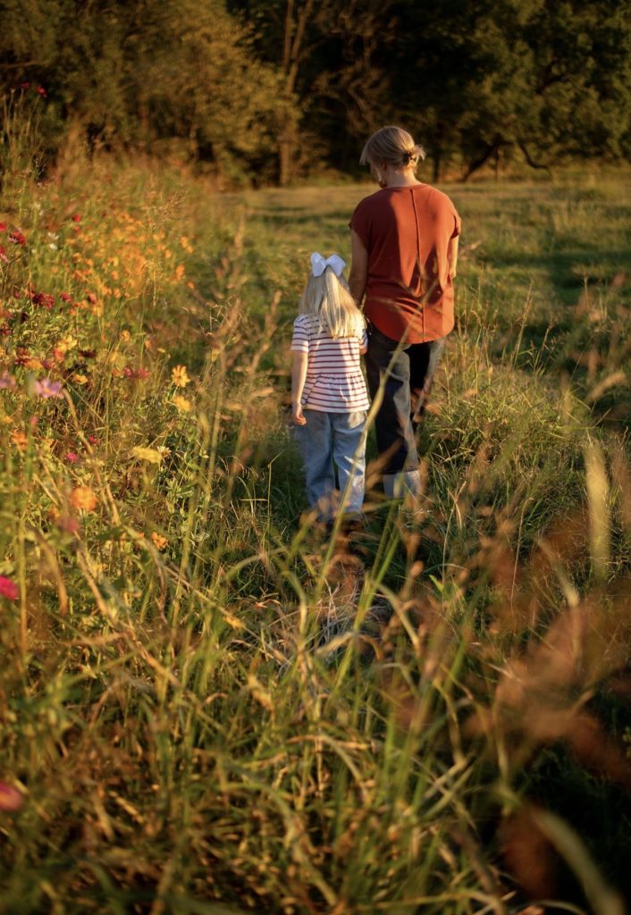 An adult and a child walking together along a wildflower path in golden light.